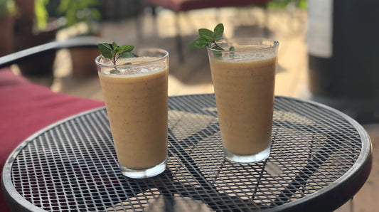 Two glasses of peach lassi garnished with fresh mint, served on a black patio table in natural sunlight