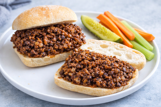 Gluten-free barbecue lentil sliders on ciabatta-style rolls with lentil quinoa filling, served with carrot sticks, celery sticks, and a dill pickle spear on a white plate