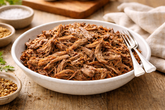 Shredded slow-cooked beef piled in a large white ceramic bowl on a wooden surface in a bright, warm kitchen setting, showing moist tender strands