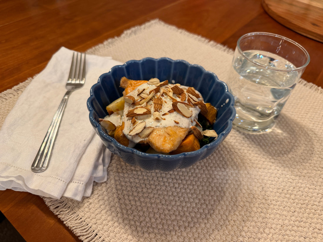 Salmon bowl with massaged kale, roasted sweet potatoes, creamy Dilliscious sauce, and sliced almonds served in a blue bowl on a tan placemat with a water glass and a fork on a cloth napkin